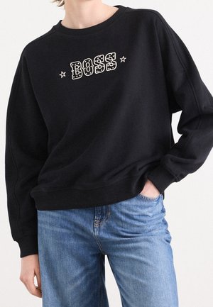 Personne portant un sweat-shirt noir avec "BOSS" et des étoiles imprimées sur la poitrine, associé à un jean bleu clair. Une main dans la poche.