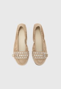 Μπεζ σουέτ φλατ παπούτσια με ελαστικό άνοιγμα και διακοσμητικό λουράκι με καρφίτσες σε ασημί τόνο και μικρή αγκράφα. Μαλακή υφή, στρογγυλεμένο σχήμα.