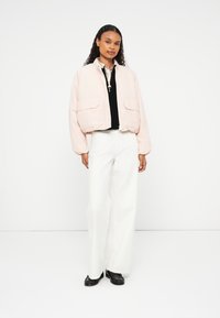 Veste cropped rose clair avec deux poches avant, poignets côtelés et fermeture éclair. Portée sur un top noir et associée à un pantalon large blanc.