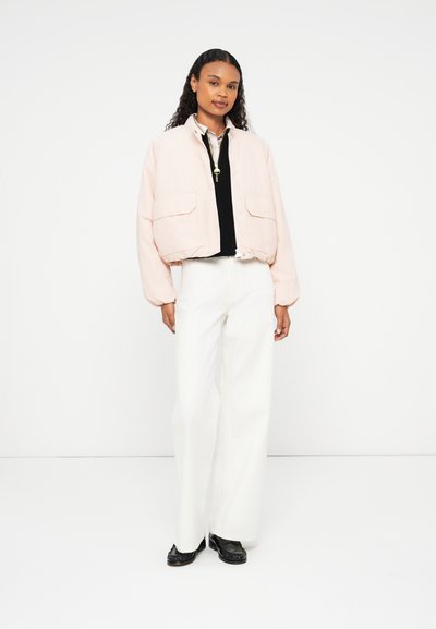 Veste cropped rose clair avec deux poches avant, poignets côtelés et fermeture éclair. Portée sur un top noir et associée à un pantalon large blanc.