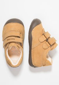 Scarpe per bambini in pelle color cuoio con suole in gomma marrone, doppie cinghie con chiusura a strappo e dettagli in cucitura bianca. Design con punta arrotondata.