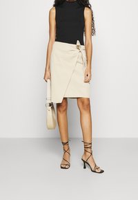 Jupe portefeuille beige avec une attache latérale, associée à un haut sans manches noir et des talons noirs à lanières. Le sac est beige clair avec une poignée circulaire.