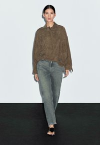 Camicia a maniche lunghe in suede marrone con una vestibilità rilassata, abbinata a jeans in denim blu sbiadito. Il modello indossa sandali neri senza fibbia. Sfondo semplice.