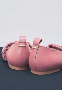 Roze leren ballerina's met een geplooide accent bij de hiel, kleine lusjes om aan te trekken en een lichtbruine zool. Soepele textuur met subtiele stiknaden.