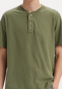 Olivovo zelené tričko s krátkym rukávom typu henley vyrobené z mäkkého materiálu. Má okrúhly výstrih s manželkou na štyri gombíky a voľný strih.