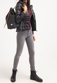 Veste matelassée noire, haut bordeaux, jean skinny gris, bottes noires à lacets et sac à dos noir à cordon avec accents marron.