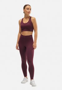 Sujetador deportivo color burdeos con escote redondeado y detalle recortado, combinado con leggings de talle alto a juego. Tejido suave y elástico, con un marcado mínimo.