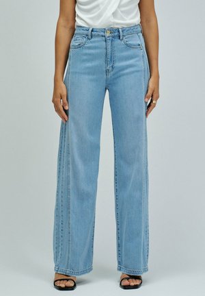 Persona con vaqueros anchos azul claro con detalles en la costura lateral, una camiseta blanca metida por dentro, y sandalias negras abiertas con tacones cuadrados.