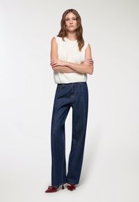 Biały, dzianinowy sweter bez rękawów z wzorem w warkocze, w połączeniu z ciemnoniebieskimi spodniami o szerokich nogawkach. Modelka ma na sobie błyszczące burgundowe szpilki.