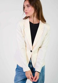 Blazer crème clair avec revers en pointe, un bouton noir, coupe ajustée, porté sur un top noir et associé à un jeans bleu.