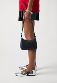 Sac en cuir noir avec une forme arrondie, quincaillerie dorée et une courte bandoulière. Porté avec un pull en tricot rouge et une jupe plissée noire.