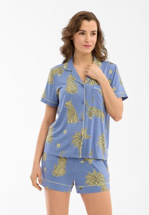 Kvinde iført blå kortærmet skjorte med knapper og shorts med gule leopard- og tropiske plante-mønstre, stående foran en ensartet hvid baggrund.