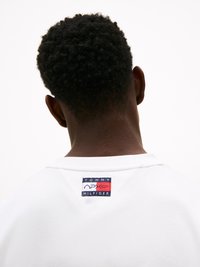 Balta sportinė maikutė su mažu raudonu, baltu ir mėlynu logotipo audiniu ant nugaros. Medžiaga turi lygią tekstūrą ir laisvą prigludimą.