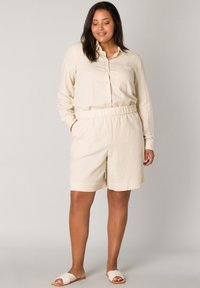 Beige linnen overhemd met knopen en een borstzak, gecombineerd met bijpassende shorts met elastische tailleband. Eenvoudig, casual ontwerp; platte sandalen in een lichte kleur.