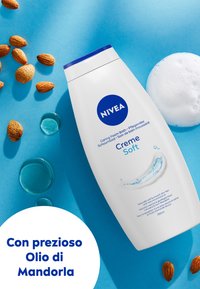 Bottiglia di schiuma da bagno NIVEA Creme Soft, bianca con tappo blu, caratterizzata da un design a onde e gocce blu. Circondata da mandorle su uno sfondo blu.