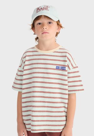 Junge mit roten Haaren, der ein weißes und rot gestreiftes T-Shirt mit dem Schriftzug "BOBO CHOSES" und eine weiße Baseballkappe mit rotem "Pickles"-Logo trägt.