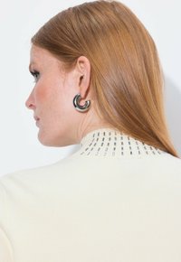 Beżowy sweter z metalicznymi akcentami na prążkowanym kołnierzu oraz błyszczącymi czarnymi okrągłymi kolczykami. Gładka faktura i nowoczesny design.