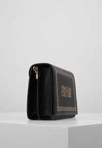 Portafoglio nero in jeans couture di Versace con cerniera dorata e bordo decorativo cucito, appoggiato su una superficie bianca su uno sfondo grigio.