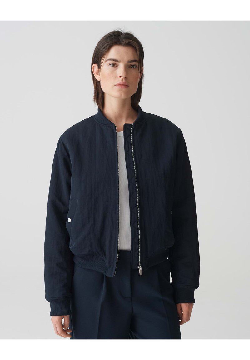 Marineblauer Bomberjacke mit gestepptem Design, vorderem Reißverschluss, gerippten Bündchen und seitlichen Taschen. Glatte Textur, lässige Passform und minimalistischer Stil.