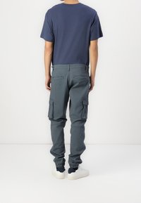 Mężczyzna w granatowej koszulce, szarych spodniach cargo z bocznymi kieszeniami i białych sneakersach, stoi przodem do białej ściany.