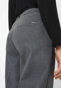 Grijze textuurbroek met een subtiel ruitpatroon, voorzien van een achterzak en een metalen logoaccent. De stof lijkt glad en aansluitend.