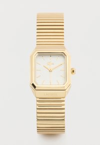 Ορθογώνιο χρυσό ρολόι χειρός με ραβδωτό μεταλλικό λουράκι, ελάχιστους χρυσούς δείκτες ώρας και λογότυπο Lacoste σε λευκό καντράν και λουράκι.