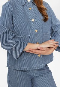 Blårandig jacka med gyllene knappar, bröstfickor och vida ärmar, tillverkad av textil med struktur, matchad med randiga byxor.