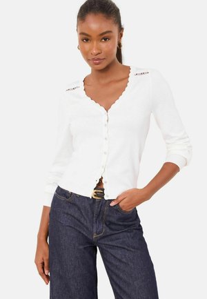 White cardigan with a scalloped neckline, button front, and lace detailing on the shoulders, paired with high-waisted dark denim pants.