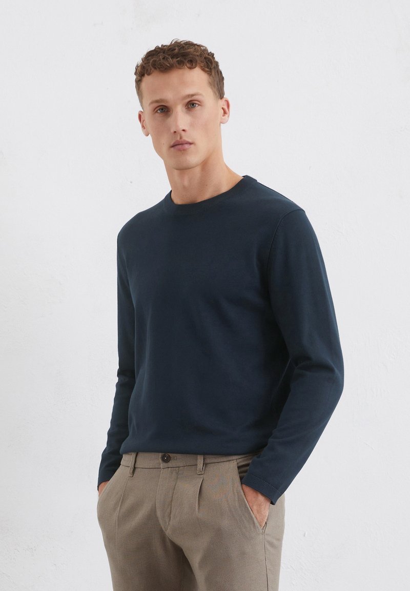 Dunkelblauses Langarm-Crewneck-Shirt aus weicher Baumwolle, mit glatter Textur und körperbetontem Schnitt, kombiniert mit beigen, plissierten Hosen.
