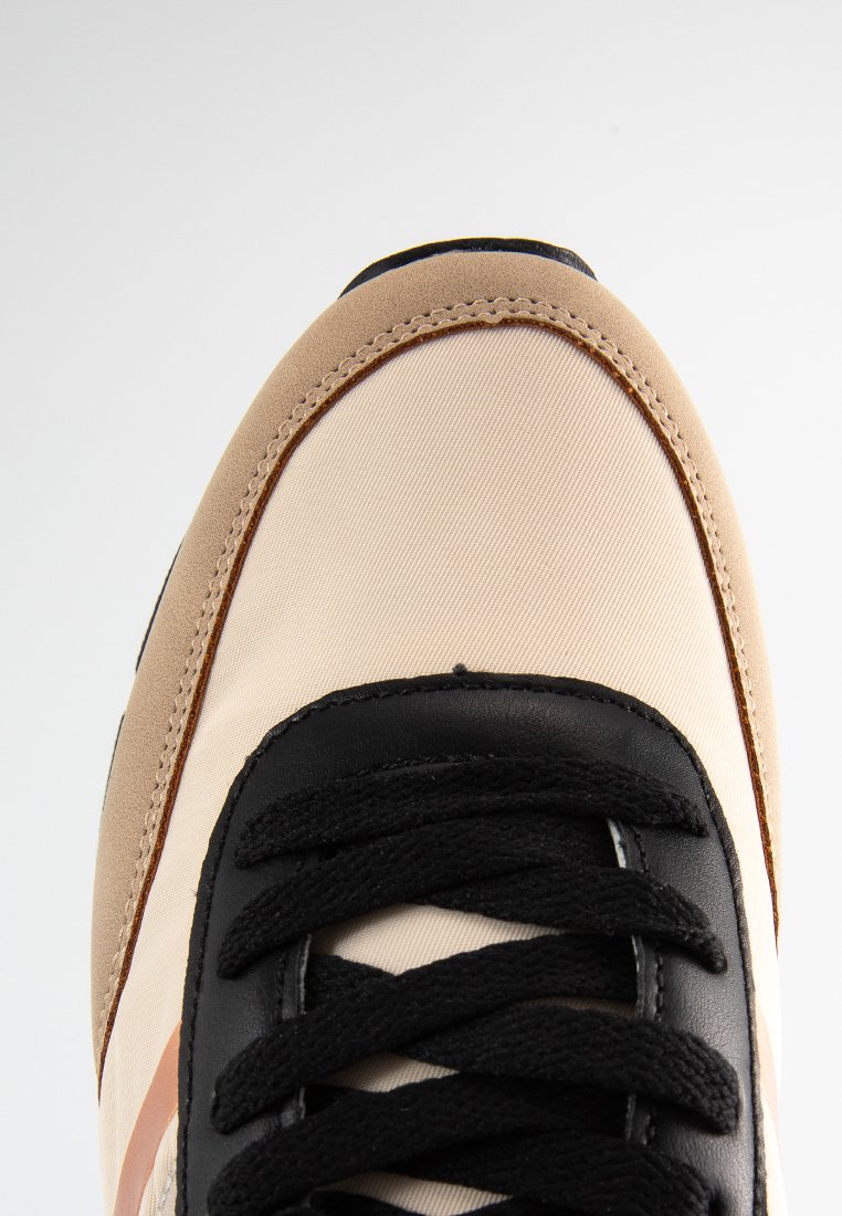 Zapatilla beige y negra con un upper de tela texturizada, cordones negros prominentes y un acento marrón contrastante a lo largo del borde de la suela.