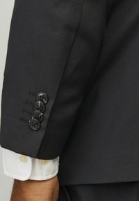 La manche d'une veste noire présente quatre boutons noirs, des coutures visibles et un poignet avec un détail en tissu blanc et un bouton.