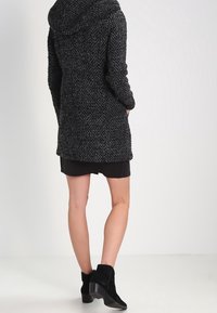 Texturerad svart-grå kappa med huva, med långa ärmar och knälång design, matchad med en svart kjol och ankelstövlar.