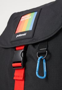 Schwarzer Ripstop-Rucksack mit einem farbenfrohen Logo-Patch, roten Riemen und einem blauen Karabinerclip. Glatte Textur mit einer strukturierten Form.