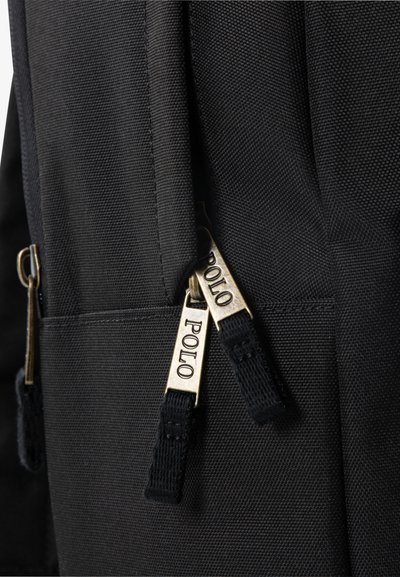 Čierny látkový batoh s dvoma zlatými zipsami a brandingom "POLO", vyrobený z textúrovaného materiálu, ktorý zdôrazňuje odolnosť a dizajn.