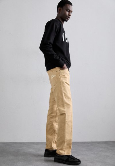 Sweatshirt noir avec motif blanc, pantalon beige avec logo sur la poche arrière, et baskets noires. Silhouette grande et fine devant un fond neutre.