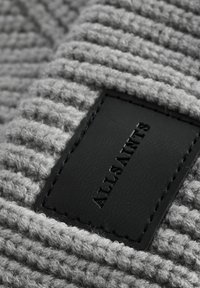 Harmaa, ribbineulottu kangas, jossa on musta nahkapatch, johon on kohokuvioitu "ALLSAINTS." Tekstuurin näyttää olevan pehmeä ja pörröinen.