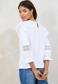 Haut en coton blanc avec des accents en dentelle sur les épaules et les manches, détail dos en clé et poignets volants. Associe à un jean en denim bleu.