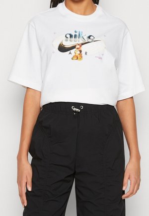Osoba v bílém tričku Nike Air s barevným grafickým motivem medvěda a černých kalhotách s nastavitelným páskem.