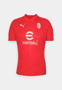 Maglia da calcio rossa con un motivo geometrico, caratterizzata da un collo a V, maniche corte e elementi di branding bianchi, inclusi logo e testo.
