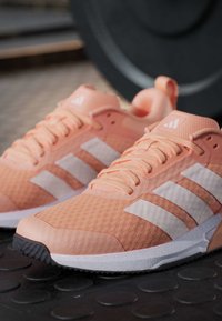 Двойка прасковени спортни обувки Adidas с бели ивици на черно спортно покритие, размит тежестен диск на заден план.