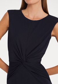 Navyblå ärmlös klänning med en twistdetalj vid midjan, slätt tyg och en rund urringning med en figurnära silhuett.