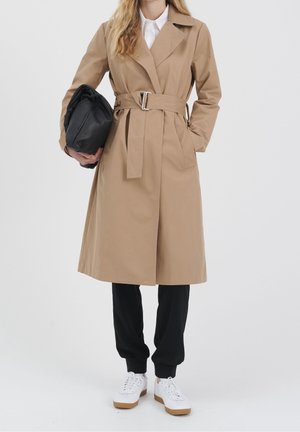 Vrouw draagt beige trenchcoat met riem, zwarte broek, witte sneakers, houdt een zwarte opgeblazen tas tegen een effen achtergrond.