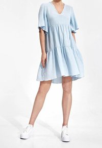 Helles blaues, gestuftes Kleid mit V-Ausschnitt und kurzen, schwingenden Ärmeln, aus strukturiertem Stoff, kombiniert mit weißen Turnschuhen.