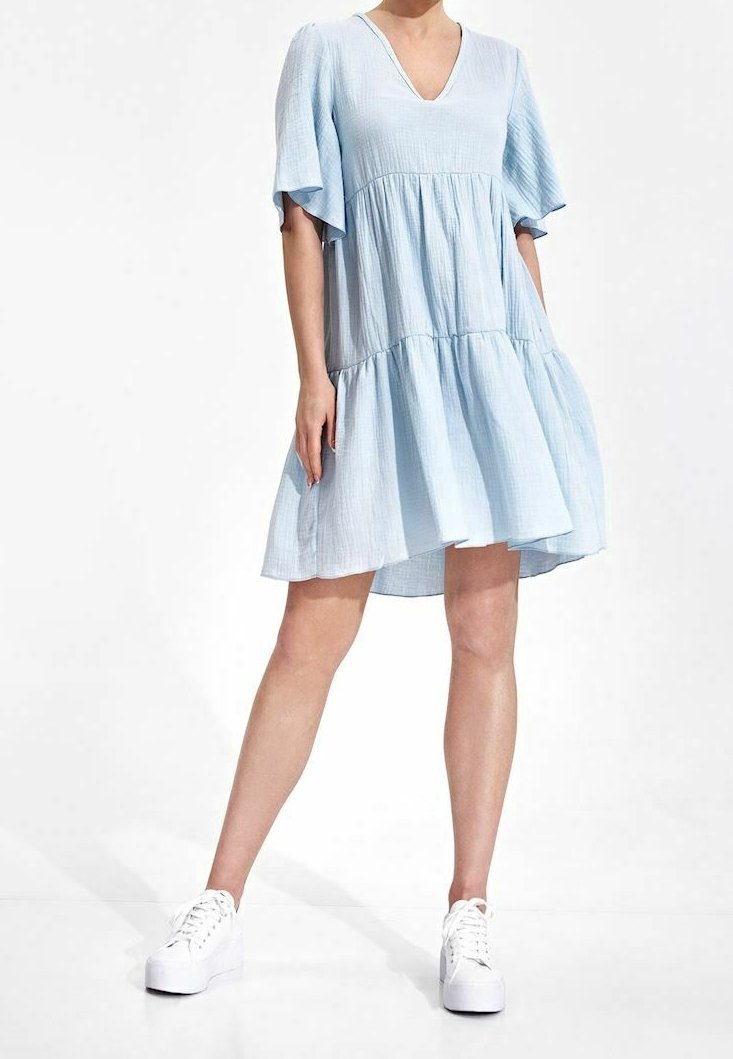 Helles blaues, gestuftes Kleid mit V-Ausschnitt und kurzen, schwingenden Ärmeln, aus strukturiertem Stoff, kombiniert mit weißen Turnschuhen.