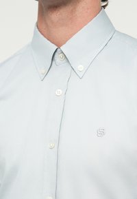 Ανοιχτό μπλε πουκάμισο με κουμπιά και γιακά με κουμπί, με μικρό κεντητό λογότυπο στο αριστερό μέρος του στήθους, φορεμένο από ένα άτομο.