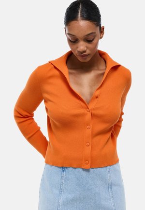 Cardigan orange côtelé avec un col évasé, présentant un devant boutonné et un ourlet court, associé à une jupe en denim clair.