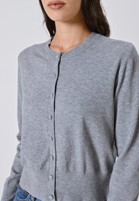 Donna che indossa un cardigan grigio con bottoni, colletto e polsini a coste, abbinato a jeans blu, su uno sfondo chiaro e semplice.