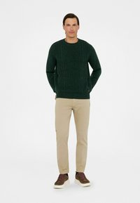 Suéter de punto trenzado verde oscuro con cuello redondo, combinado con chinos beige y zapatillas marrones con acentos texturizados y suela gruesa.