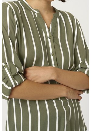 Personne portant une chemise vert olive à rayures blanches verticales, manches retroussées, bras croisés à la taille.