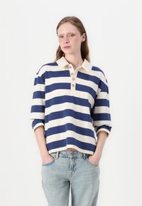 Striped polo shirt featuring wide navy and cream horizontal stripes, a collared neckline, and three-button placket. Loose fit with cuffed sleeves.